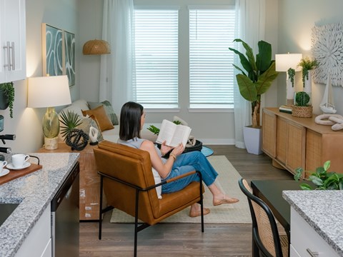 A woman is sitting on a chair reading a book in a well-lit room with a kitchen counter and various plants.