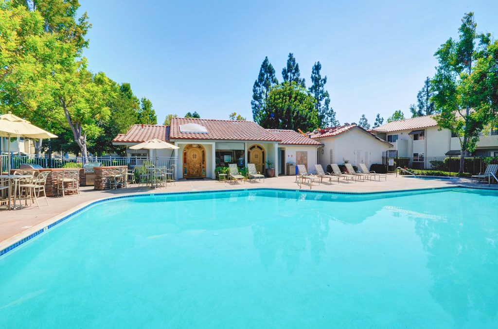 A large swimming pool in front of a house with a patio and trees in the background.