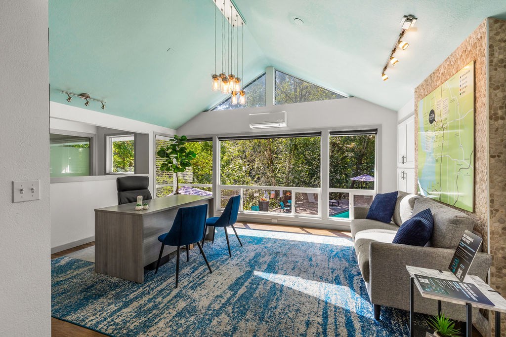 A room with a desk, chairs, and a couch with a view of trees outside the window.