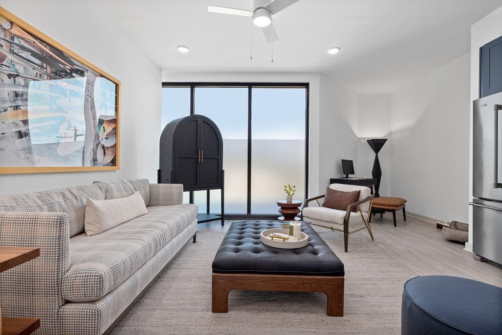 A living room with a grey couch, a black ottoman, a wooden coffee table, and a large window with a view of the ocean.