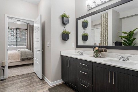 A bathroom with a large mirror, a sink, and a bedroom with a bed and window.