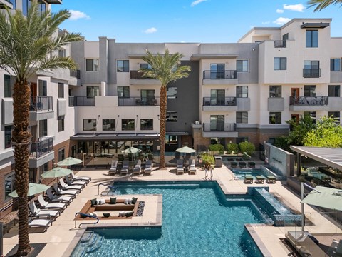 A large swimming pool surrounded by lounge chairs and umbrellas in front of a multi-story apartment building.