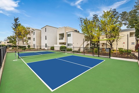 A tennis court with a blue surface and white lines is surrounded by a fence and trees.