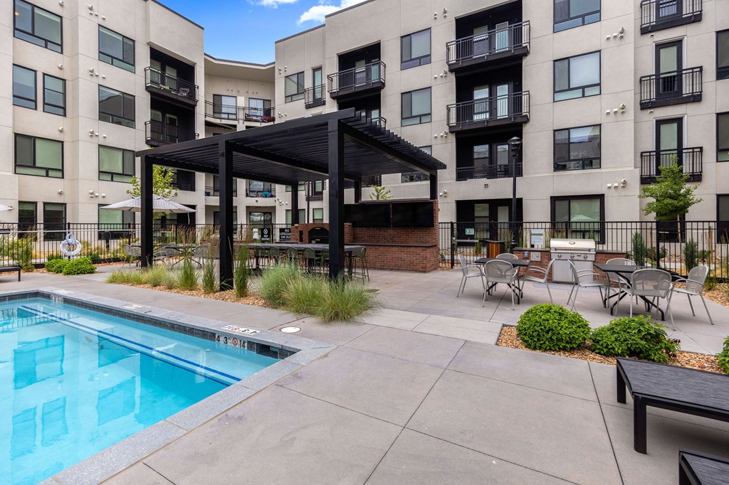 A pool area with a black pergola and chairs in front of apartment buildings.