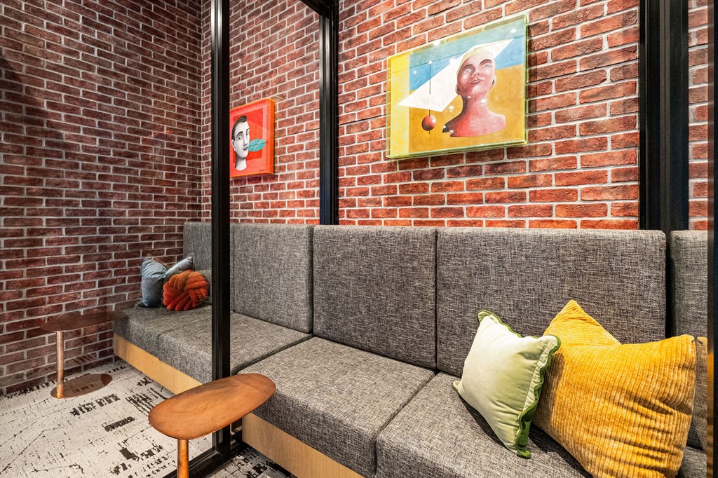A grey couch with a wooden stool in front of a brick wall with two framed pictures on it.