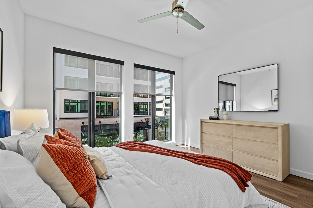 A bedroom with a large bed, a ceiling fan, and a view of a cityscape through the windows.
