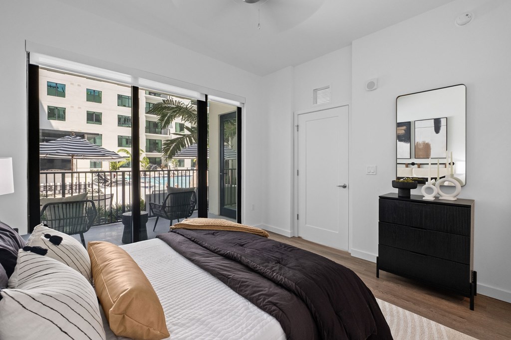 A bedroom with a large bed and a balcony with a view of a pool and buildings outside.