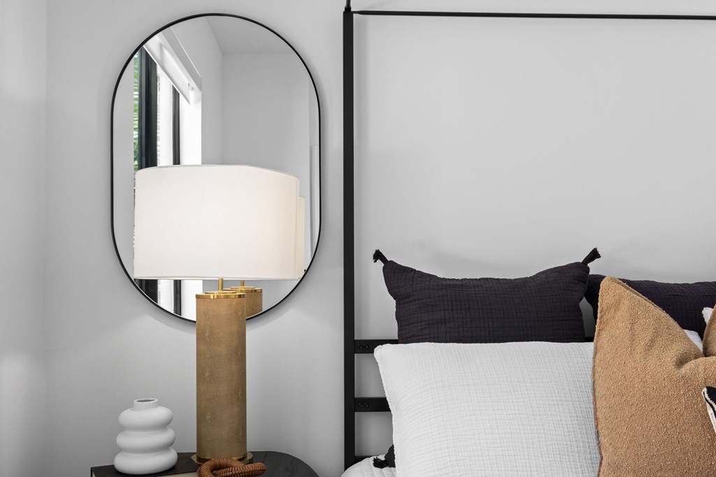 A lamp with a white shade is on a table next to a bed with pillows.