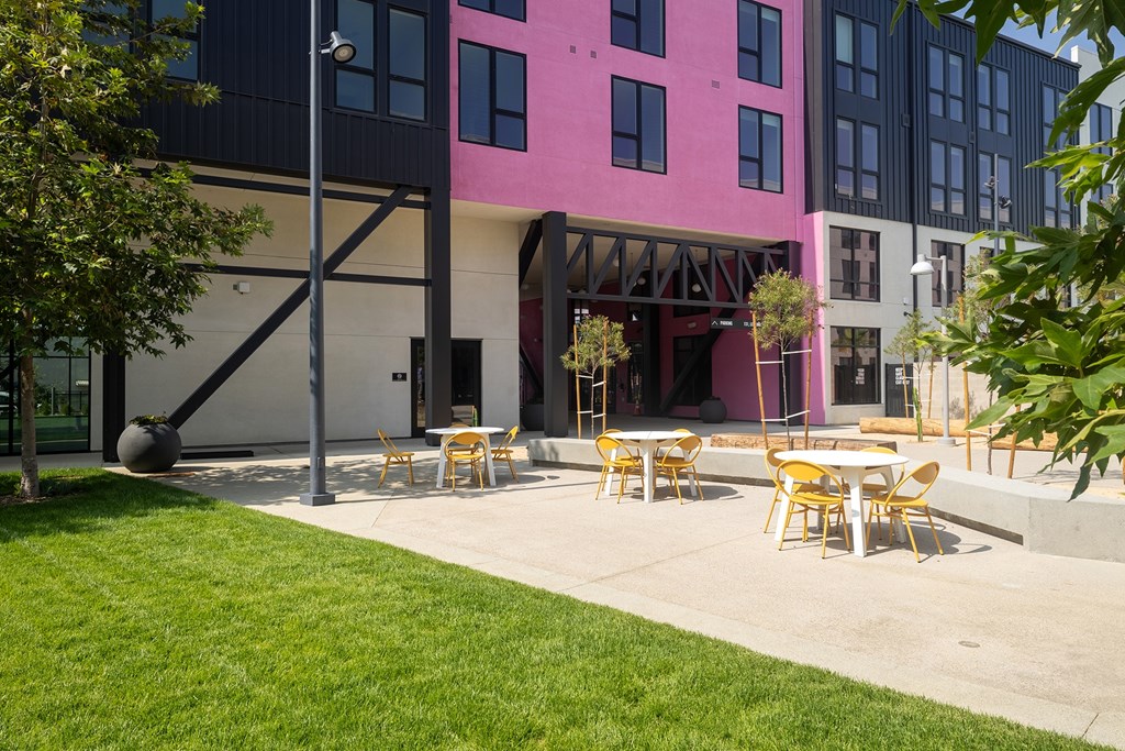 A modern building with a pink section and a patio with tables and chairs.