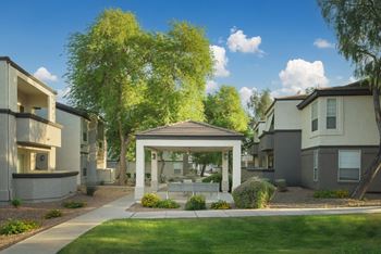 A small pavilion is in the middle of a grassy area in front of apartment buildings.