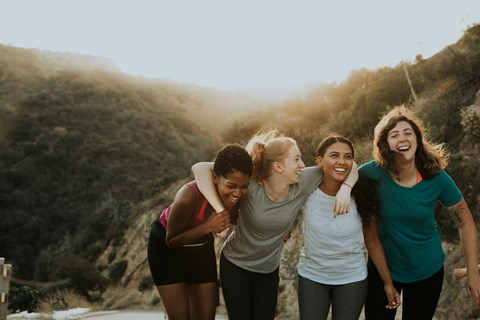 Four women are walking together on a path with a hill in the background.