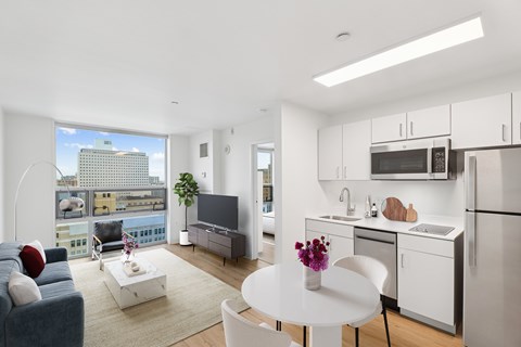 A modern kitchen with white cabinets and appliances, a dining table with chairs, and a living room with a sofa and a coffee table.