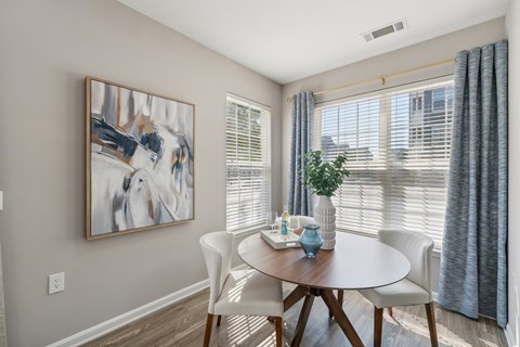 A dining room with a white table and chairs, a painting on the wall, and a window with blinds.