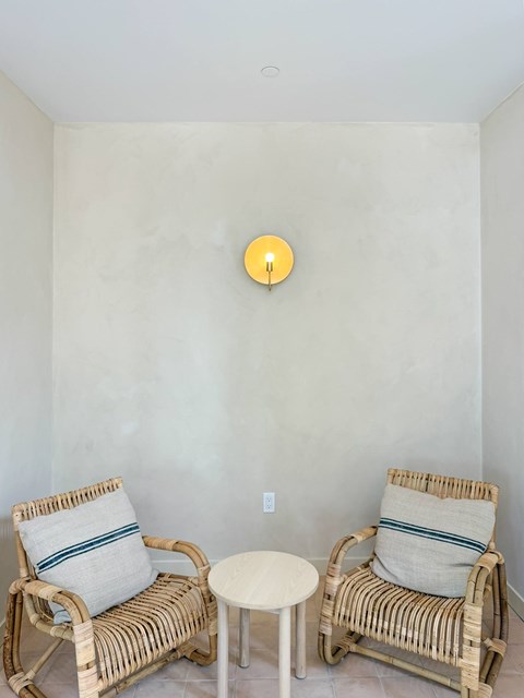 Two wicker chairs with cushions and a small table are placed in a room with a wall clock.