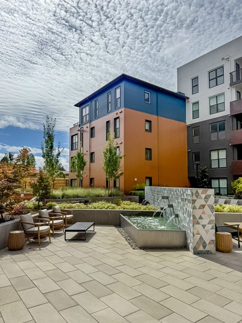 A modern building with a blue, orange, and pink facade is surrounded by a courtyard with a fountain and seating area.