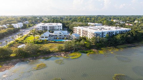 A large white building with a pool in front of it is surrounded by greenery and water.