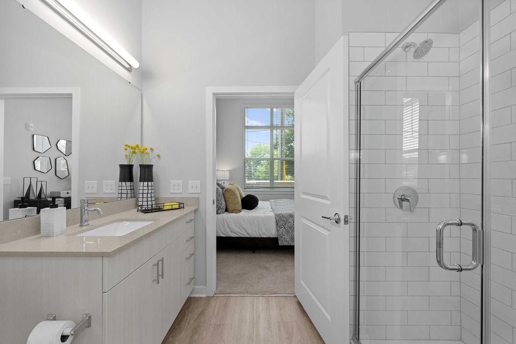 A white bathroom with a walk-in shower and a bed in the background.