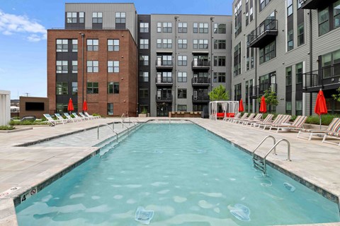 A swimming pool surrounded by chairs and umbrellas in front of apartment buildings.