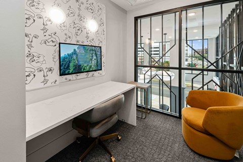 A white desk with a grey chair and a yellow armchair in front of a wall with a TV screen and a window.