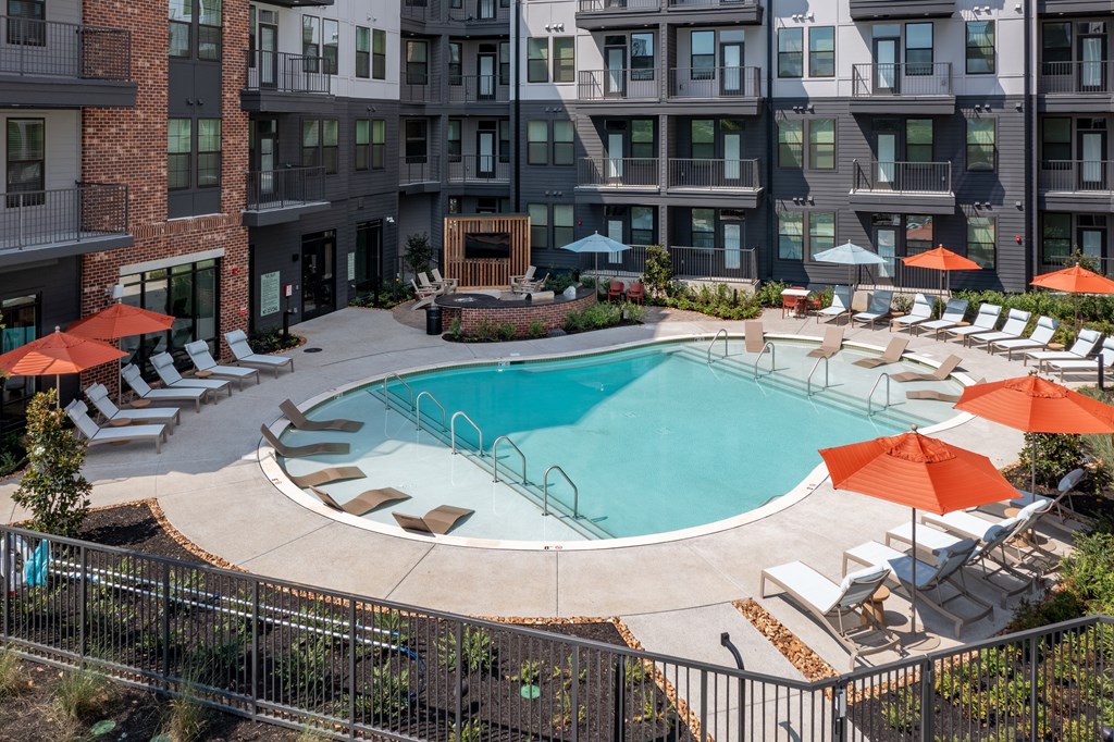 A large swimming pool surrounded by lounge chairs and umbrellas in front of a multi-story apartment building.