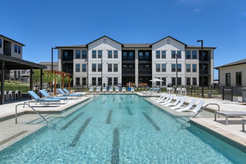 A large swimming pool in front of a multi-story building with lounge chairs around it.