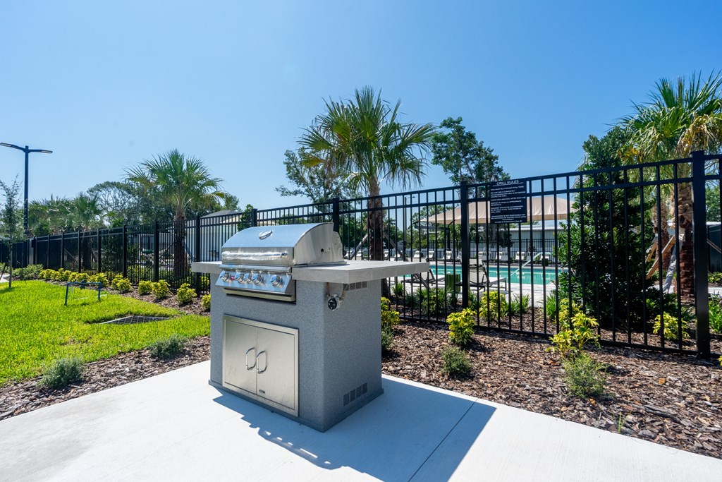 A grey trash bin with a lid and a handle is placed on a concrete surface surrounded by a black fence and greenery.