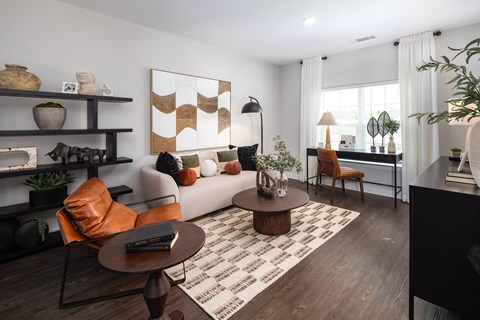A living room with a brown and white rug, a brown coffee table, a white sofa, a brown chair, a black bookshelf, and a painting on the wall.