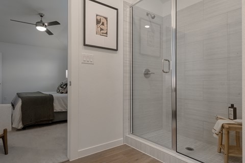 A bathroom with a glass shower door and a bedroom with a ceiling fan.