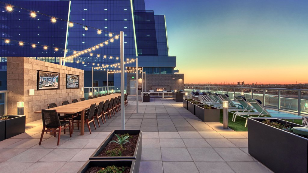 A rooftop patio with a long table and chairs, string lights, and a view of the city skyline at dusk.