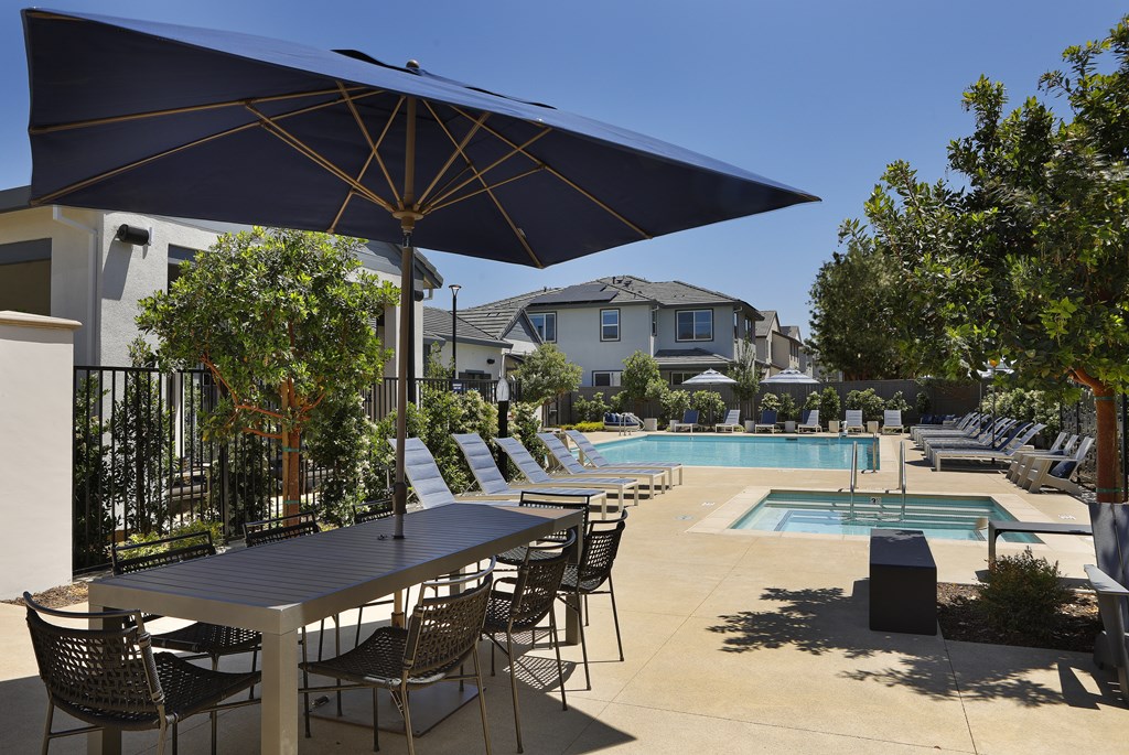 A table with chairs and an umbrella is in the foreground of a pool area.