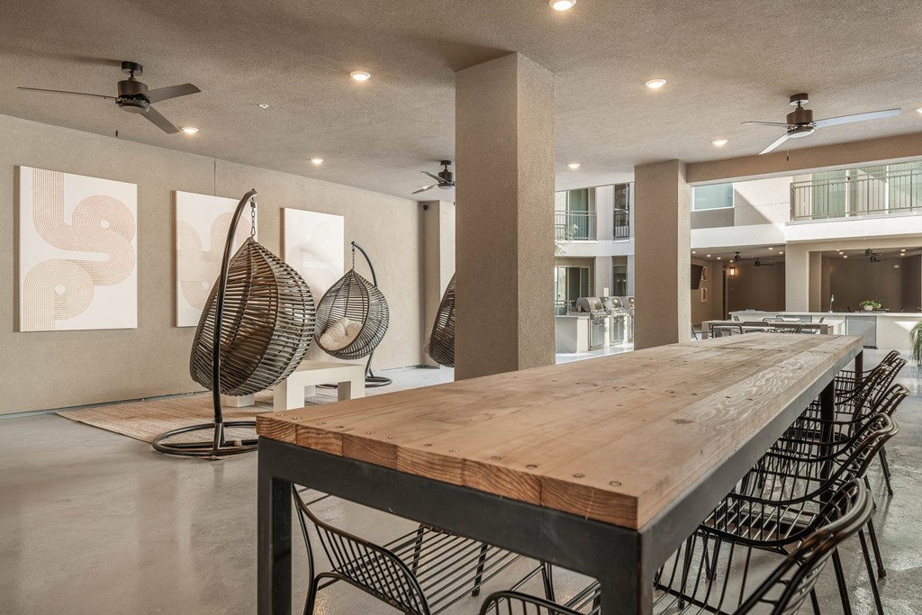 A long wooden table with chairs is in the foreground of a spacious room with a fan and artwork on the wall.
