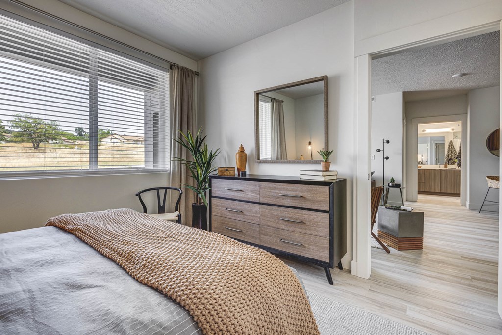 Angled bedroom shot with dresser, mirror, bed, and window with open door to living area