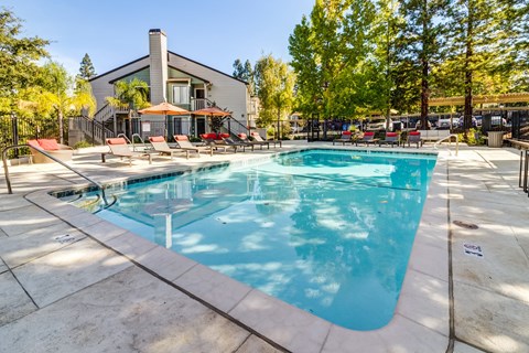 A large outdoor swimming pool surrounded by a patio area with chairs and a building in the background.