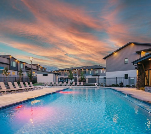 A swimming pool surrounded by lounge chairs and buildings under a colorful sky.