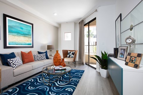 A living room with a grey couch, a blue patterned rug, and a painting of a beach scene on the wall.