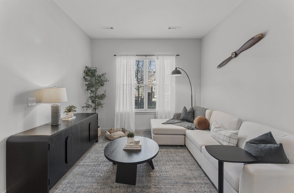 A modern living room with a white sofa, a black coffee table, and a large window with white curtains.