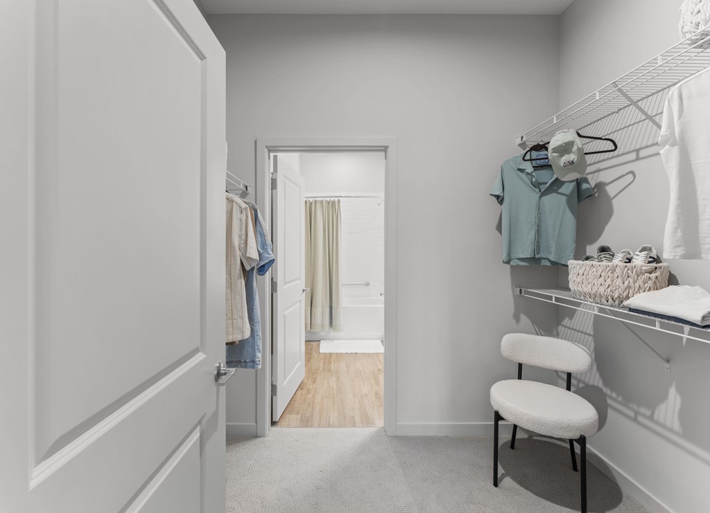 A white closet with clothes hanging and a chair in front of a mirror.