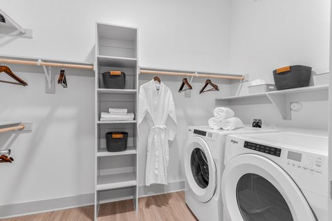 A white laundry room with a washer and dryer, a white robe hanging up, and a white shelf with a black container on it.