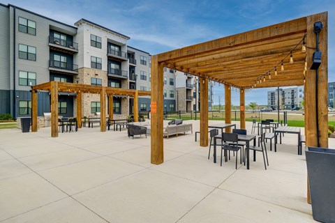 A wooden pergola is over a patio with black chairs and tables.