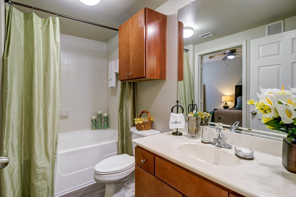 A bathroom with a white toilet, a white bathtub with a green shower curtain, a wooden medicine cabinet, a white sink with a mirror above it, and a vase of flowers on the counter.