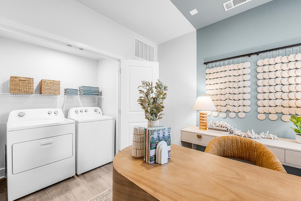 A laundry room with a washer and dryer, a table with a plant and a vase, and a window with a curtain.