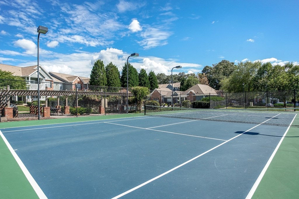 A tennis court with a blue surface and white lines, surrounded by a fence and trees.