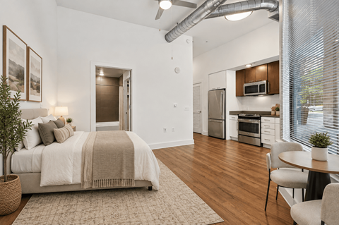 A bedroom with a bed, chair, and a kitchen area in the background.