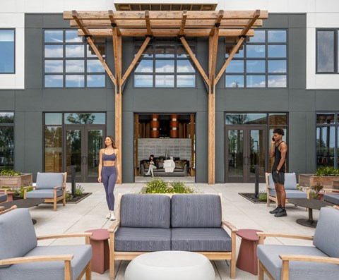 A woman in a black top and grey leggings stands in front of a fireplace in a modern building with a wooden pergola above.