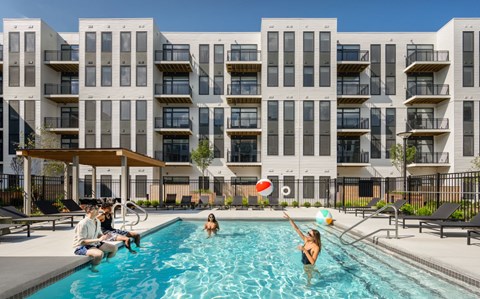 People are enjoying a sunny day at the pool in front of a modern apartment building.
