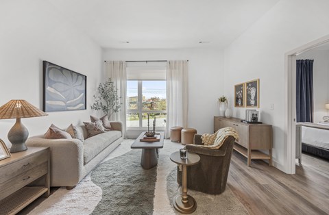 A well-lit, spacious living room with a large window, a sofa, a coffee table, and a side table with a lamp.