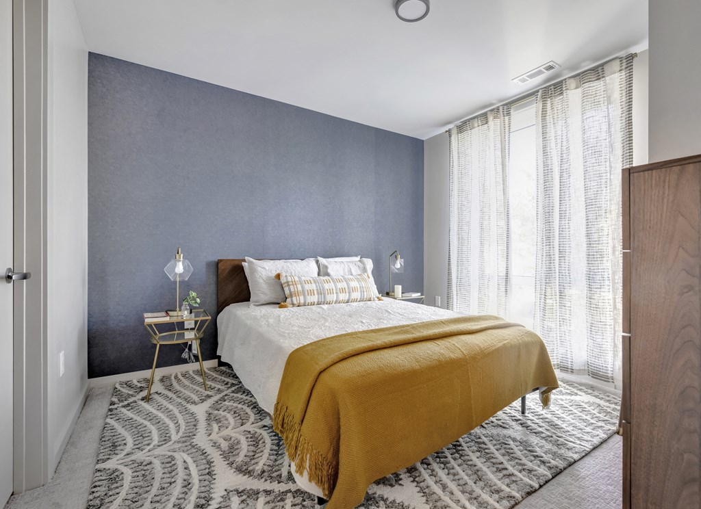 Model bedroom with floor to ceiling window, blue fabric accent wall, and modern decor