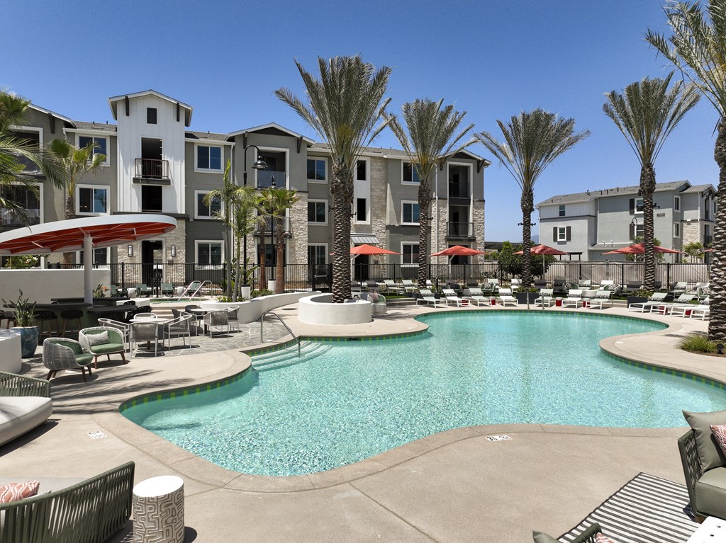 A large swimming pool surrounded by palm trees and lounge chairs in front of apartment buildings.