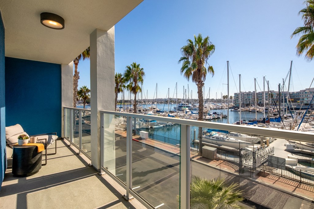 A balcony with a glass railing overlooking a marina with boats and palm trees.