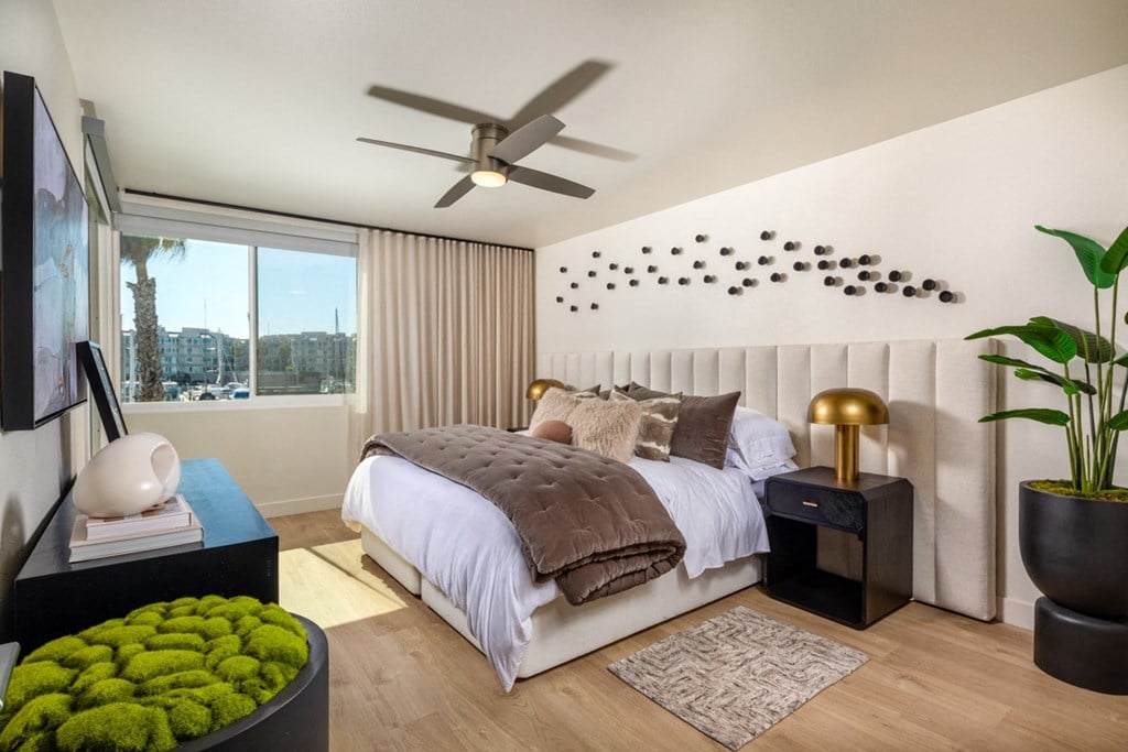 A bedroom with a bed, a fan, and a window with a view of a city.